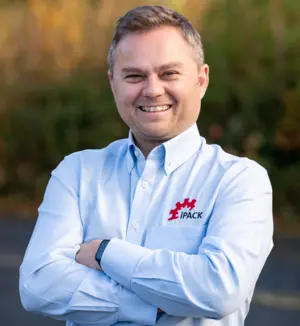 John Hall de iPack Solutions, agente de IMPACK en el Reino Unido, sonriendo con los brazos cruzados y una camisa azul claro con el logotipo de iPack Solutions.