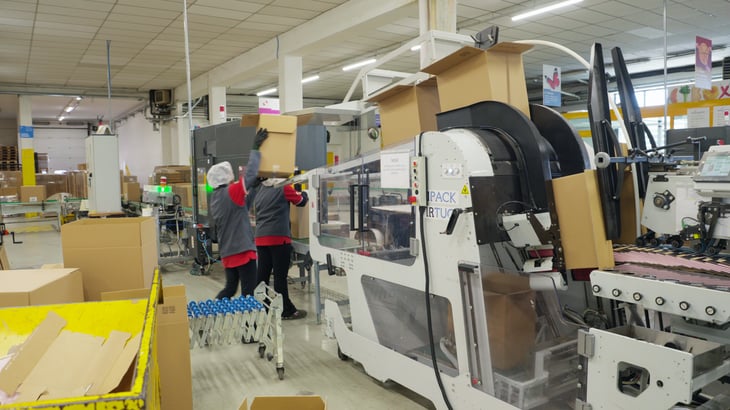 Confezionatrice automatica VIRTUO di IMPACK nel reparto di produzione di Box Marche, con due operatori che movimentano scatole di cartone nelle vicinanze.
